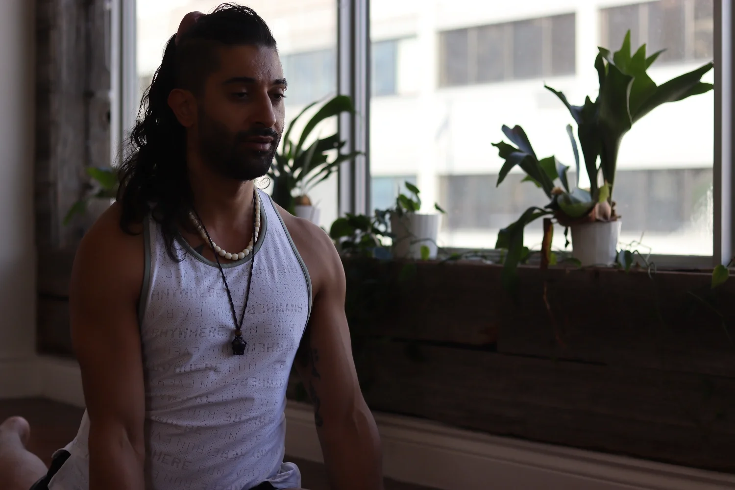 A man with long dark hair, wearing a white sleeveless shirt, pearl necklace, and a black necklace with a pendant, sitting inside near a window with potted plants on the windowsill.