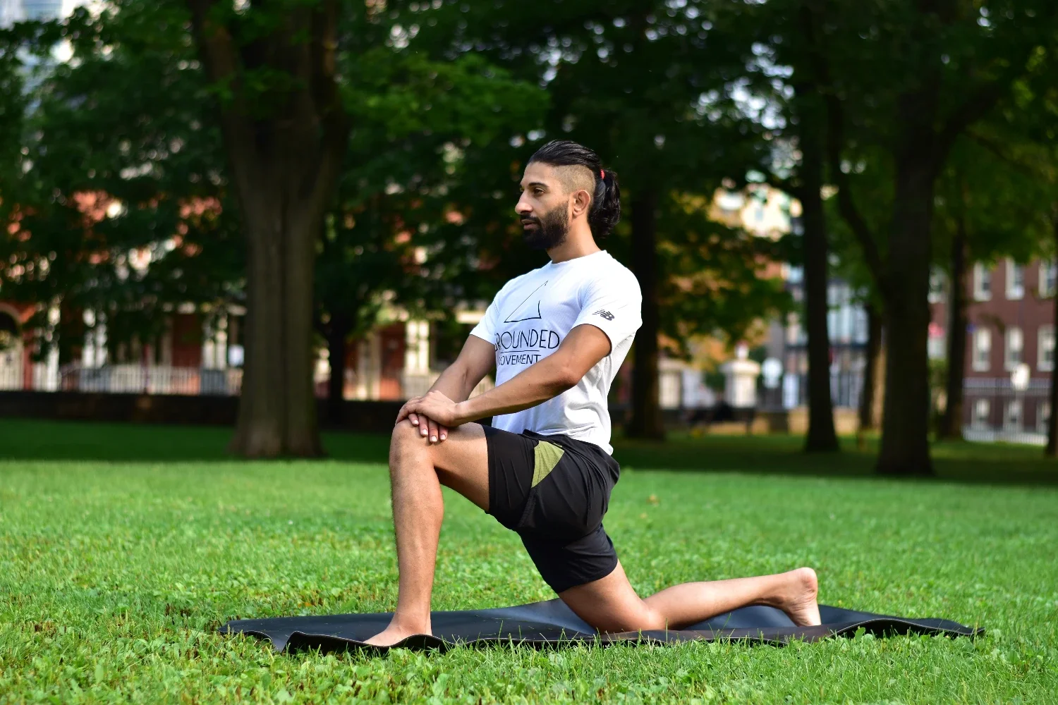 A man with a beard and long hair in a ponytail practicing yoga outdoors on a grassy field with trees and buildings in the background, kneeling with one knee bent and hands on his thigh.