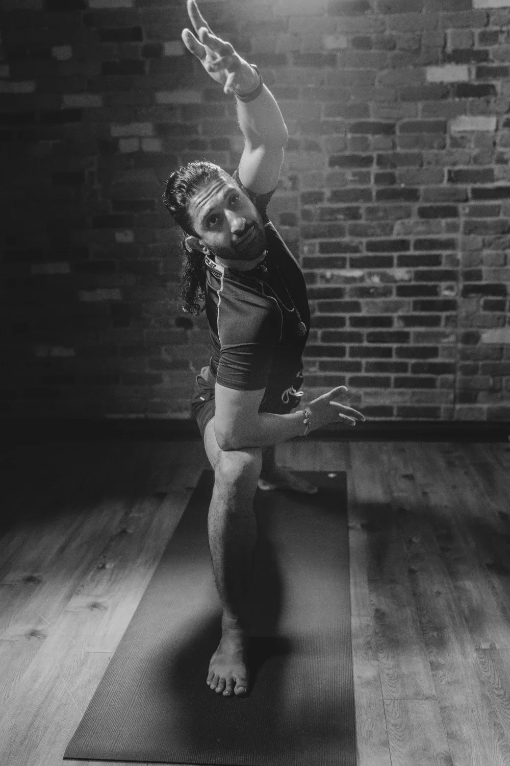 Black and white photo of a person practicing yoga on a mat against a brick wall, balancing on one leg with one arm extended upward and the other arm extended outward, looking at the camera.
