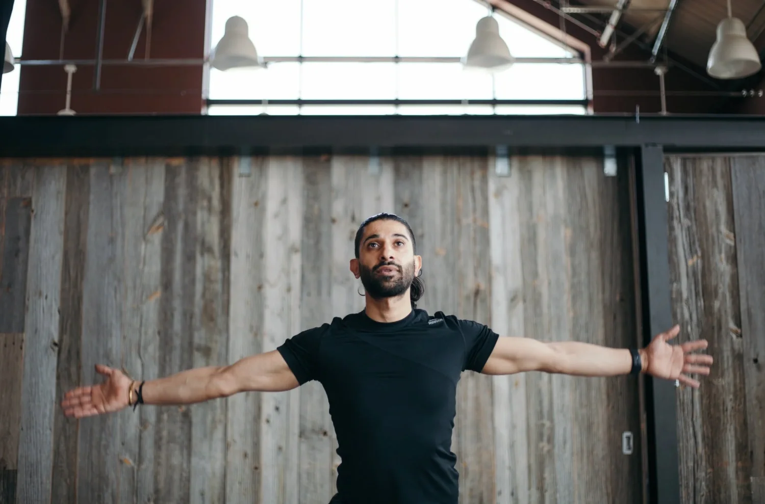 A man with a beard and black shirt standing indoors with arms extended to the sides, in front of a wooden wall, with large windows and industrial lighting above.