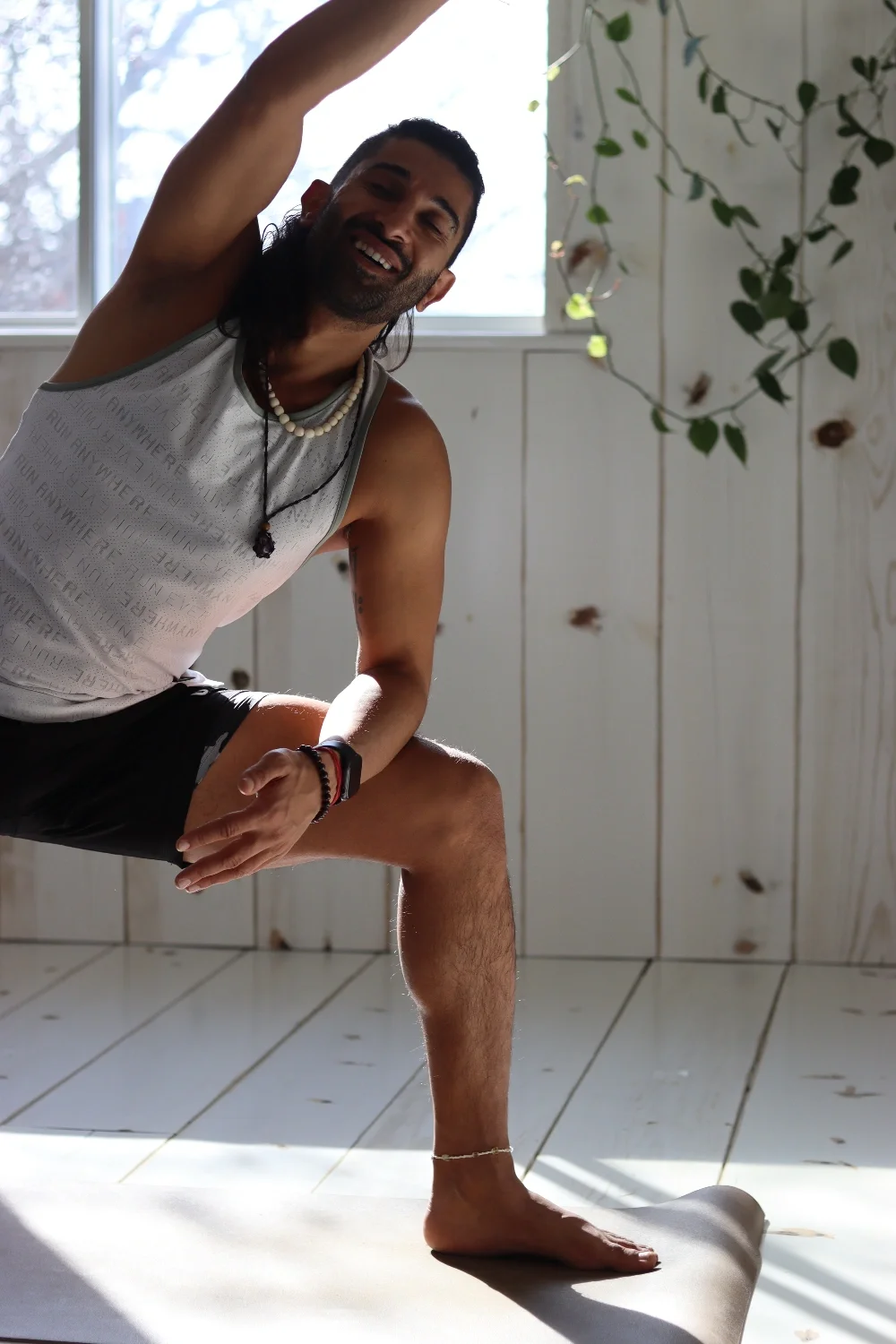 Man practicing yoga indoors, standing on one leg on a yoga mat, performing a balance pose, with a window and some greenery in the background.