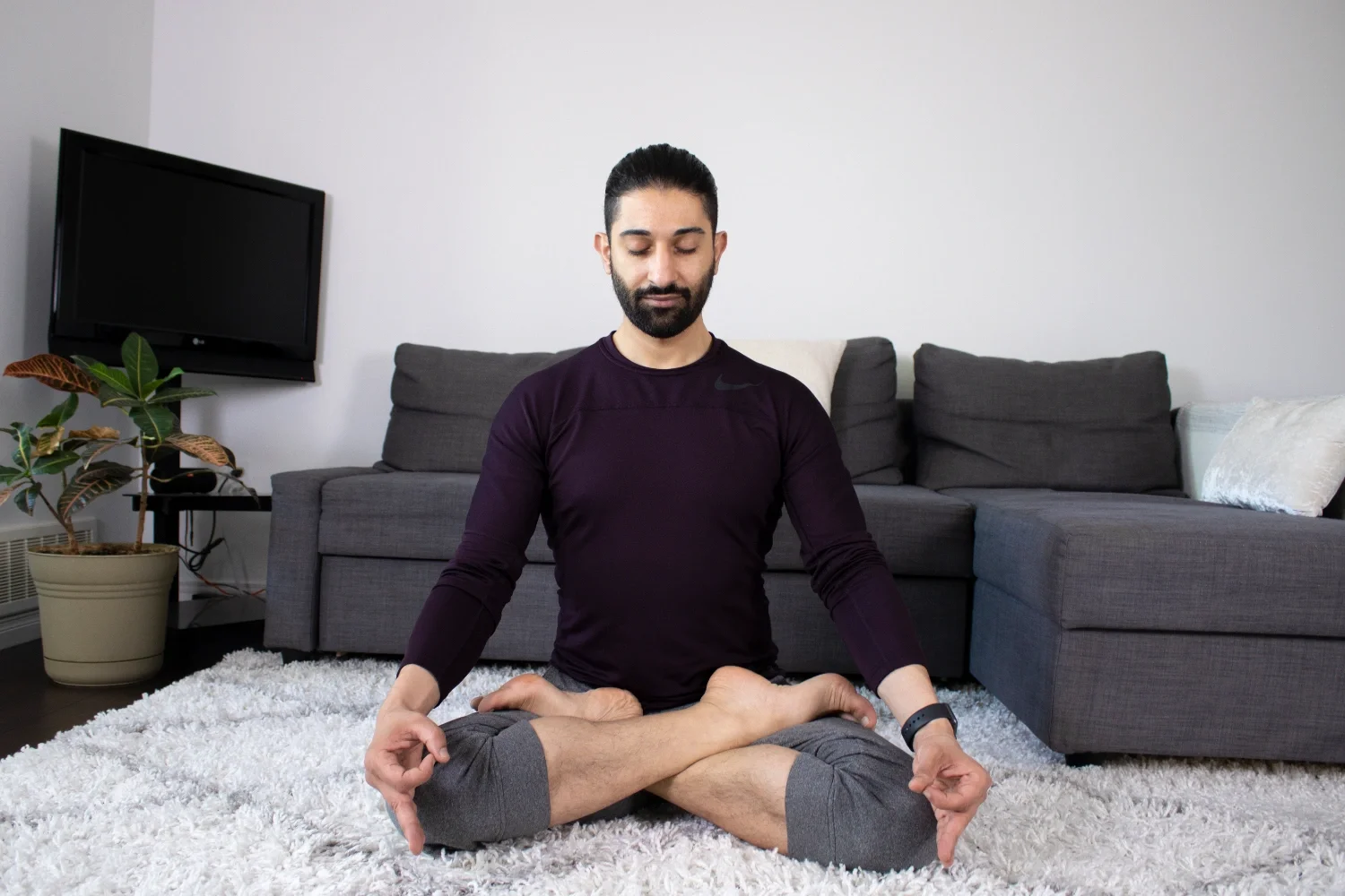 Man practicing yoga in a living room, sitting cross-legged on a white shag rug with eyes closed.