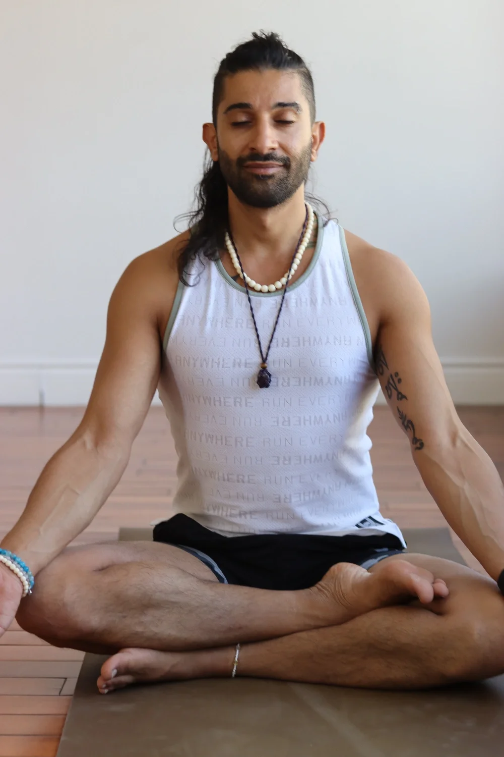 A man practicing yoga in a seated lotus position on a yoga mat, with his eyes closed and a peaceful expression, wearing a tank top, shorts, necklaces, and bracelets, indoors with a plain wall background.