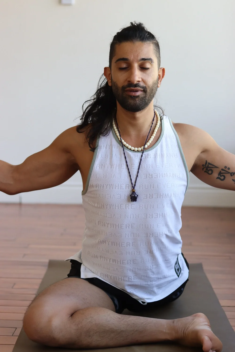 A man with long dark hair and facial hair practicing yoga indoors on a mat, sitting in a cross-legged position with closed eyes, wearing a white sleeveless shirt, black shorts, and layered necklaces.