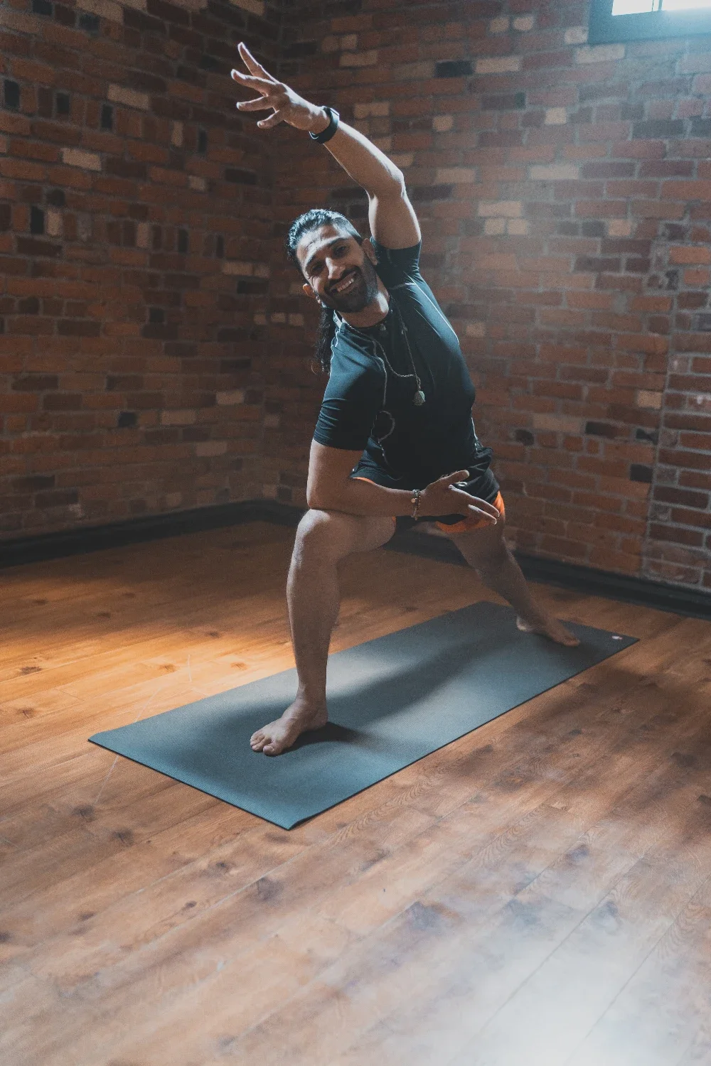 A man practicing yoga on a black mat in a room with wooden floors and brick walls, wearing a black shirt and shorts, smiling and striking a playful pose.