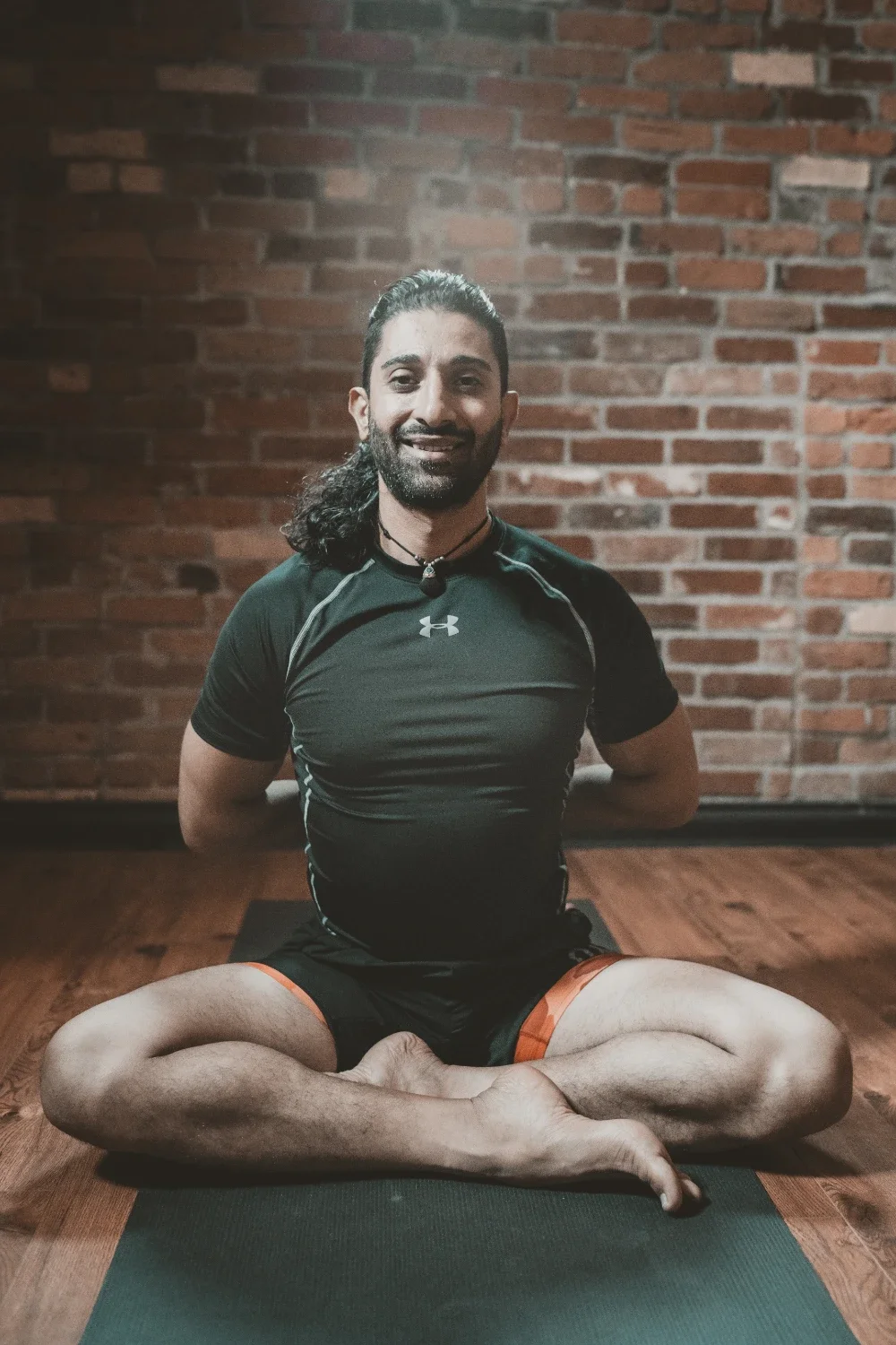 A man with long dark hair, a beard, and a choker necklace sitting cross-legged on a black yoga mat in front of a brick wall, smiling, wearing a black Under Armour athletic shirt and shorts.