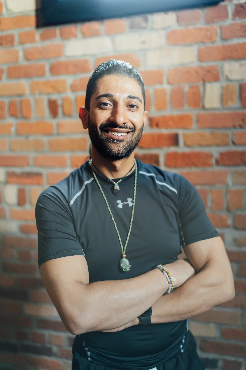 Smiling man with a beard and long hair in a black athletic shirt, standing with arms crossed in front of a brick wall.