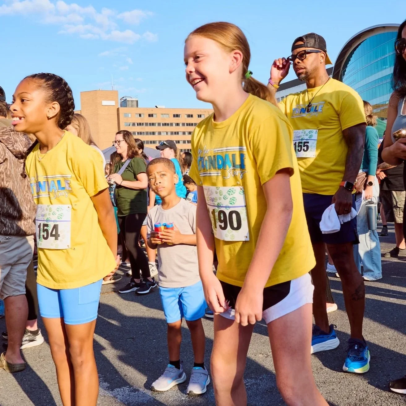 People gather outdoors for a race event, wearing yellow T-shirts with bib numbers, with a building and blue sky in the background.
