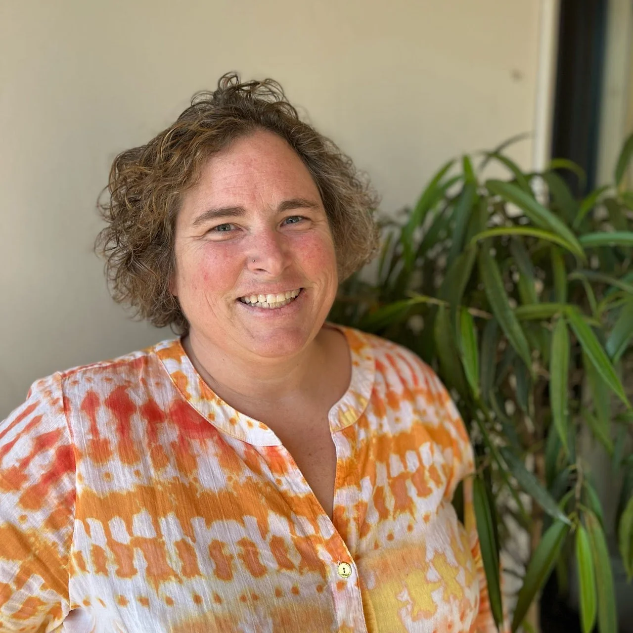 A woman smiling outdoors next to a green plant, wearing an orange and white patterned shirt.