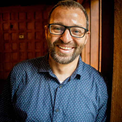A smiling man wearing glasses and a blue patterned shirt, standing indoors near a wooden wall.