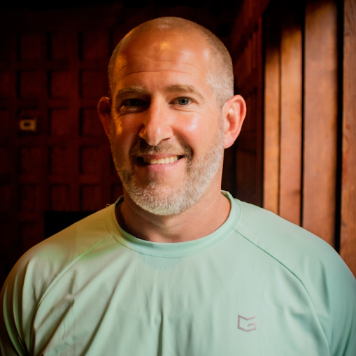 A smiling man with a shaved head and beard, wearing a light green athletic shirt, standing indoors with wooden paneling background.