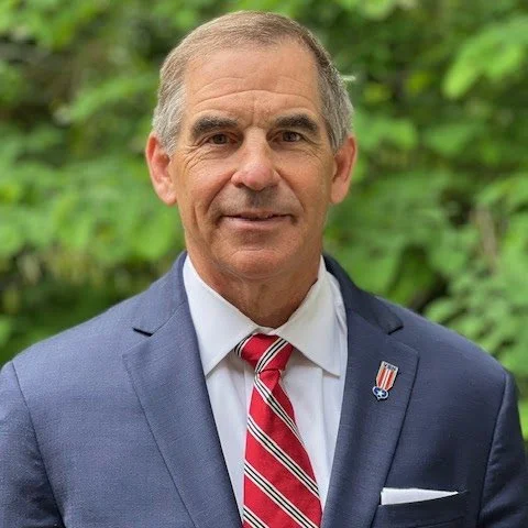 A middle-aged man in a blue suit, white shirt, and red striped tie, standing outdoors with greenery in the background.