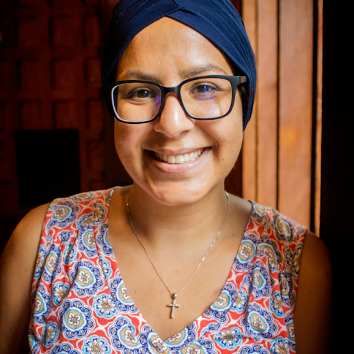 A smiling woman wearing glasses, a blue headscarf, and a patterned sleeveless top, standing in front of a wooden background.