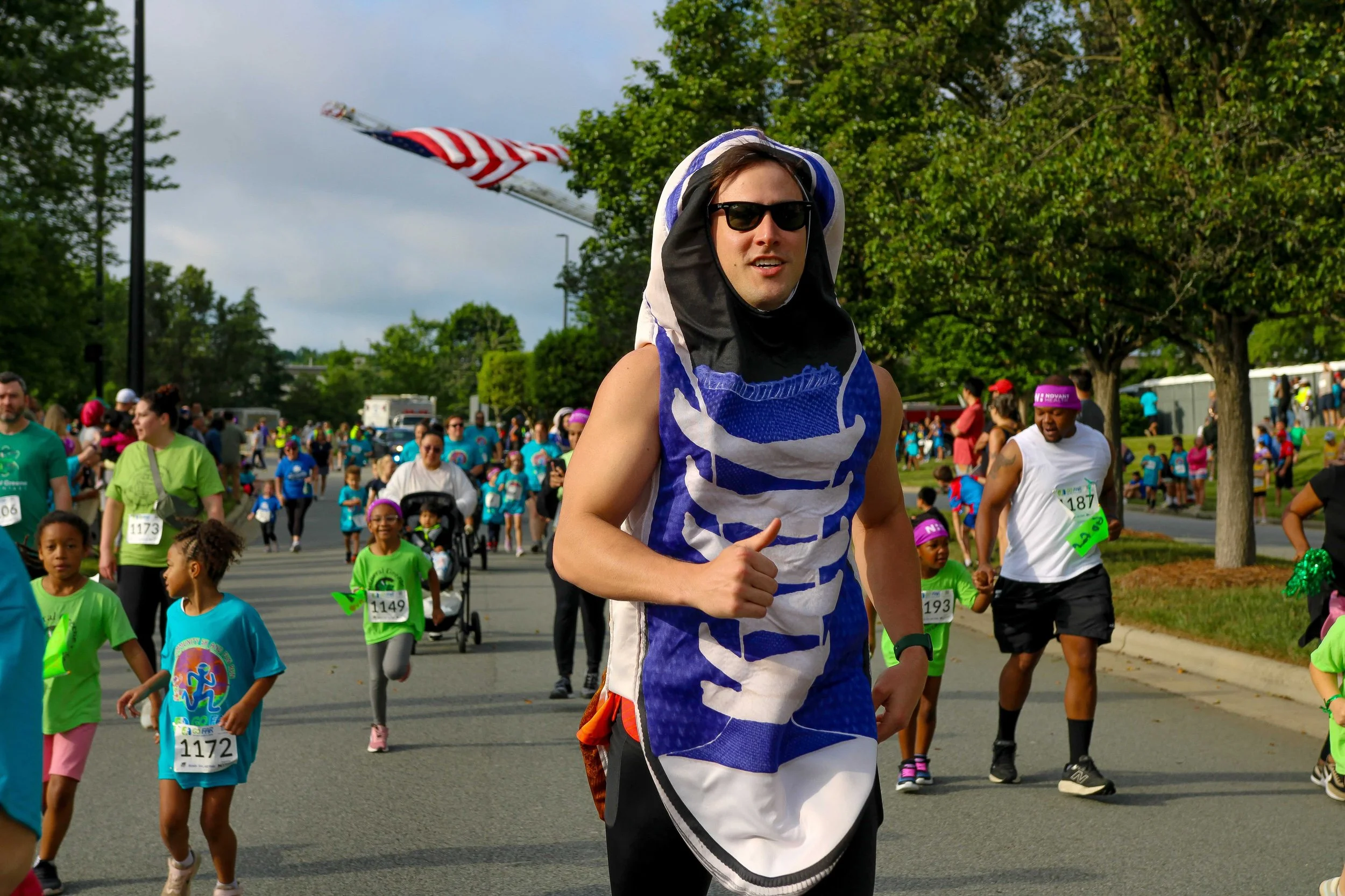 A man wearing sunglasses, a blue and white striped sleeveless shirt, and black shorts running in a community race or fun run event with a crowd of children and adults in colorful athletic clothing around him. There are trees and an American flag in the background.