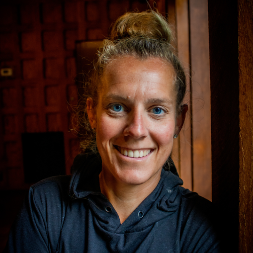 A woman with blonde hair tied in a bun, blue eyes, and wearing a dark jacket, smiling indoors with wooden walls in the background.