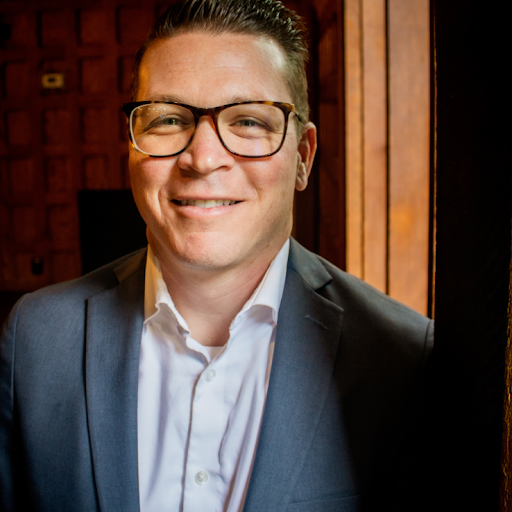 Portrait of a young man wearing glasses, a white shirt, and a dark blazer, smiling in an indoor setting with wooden background panels.