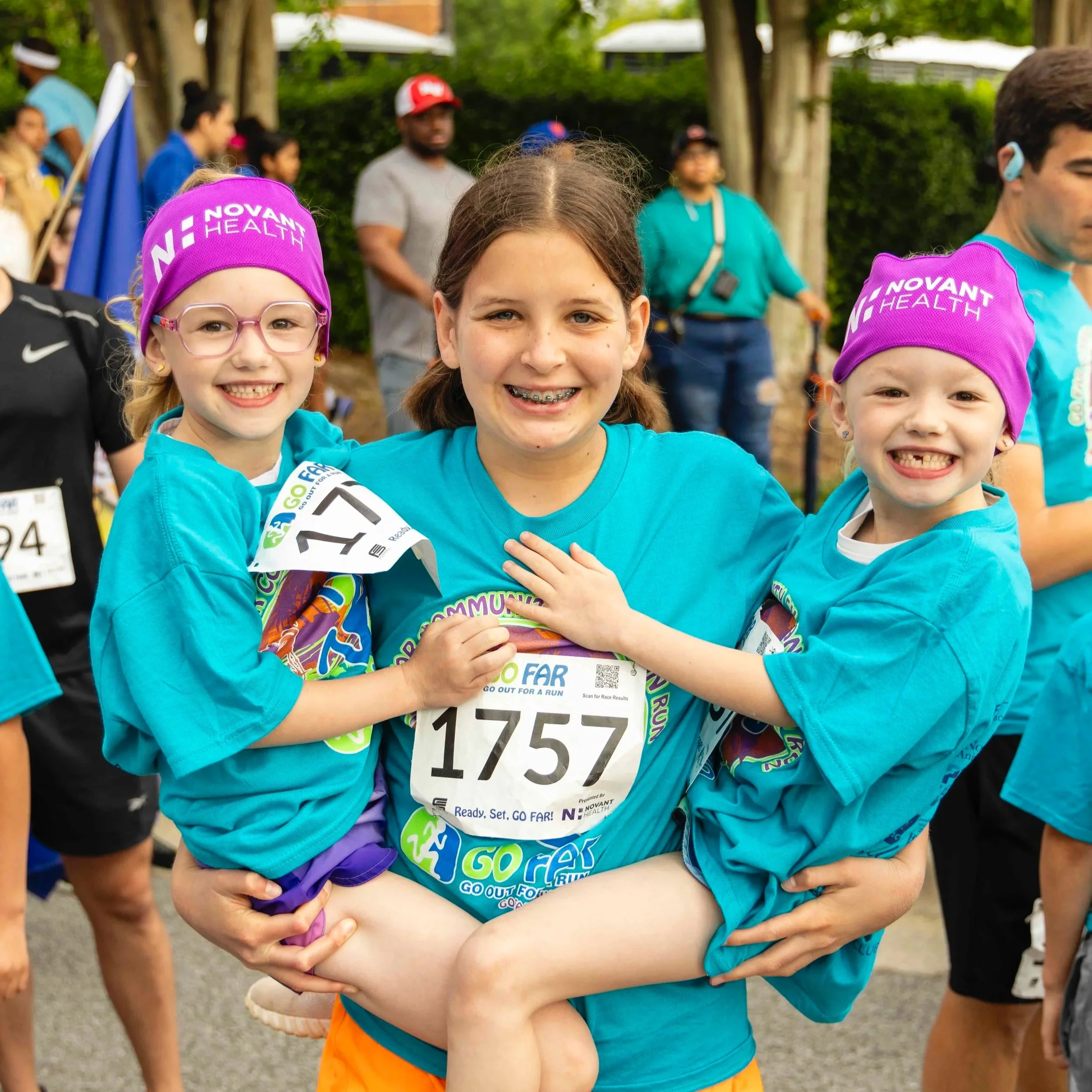 Two young girls in teal shirts and purple hats with 'NOVANT HEALTH' logos, smiling and hugging each other at a running event, with other participants in the background.