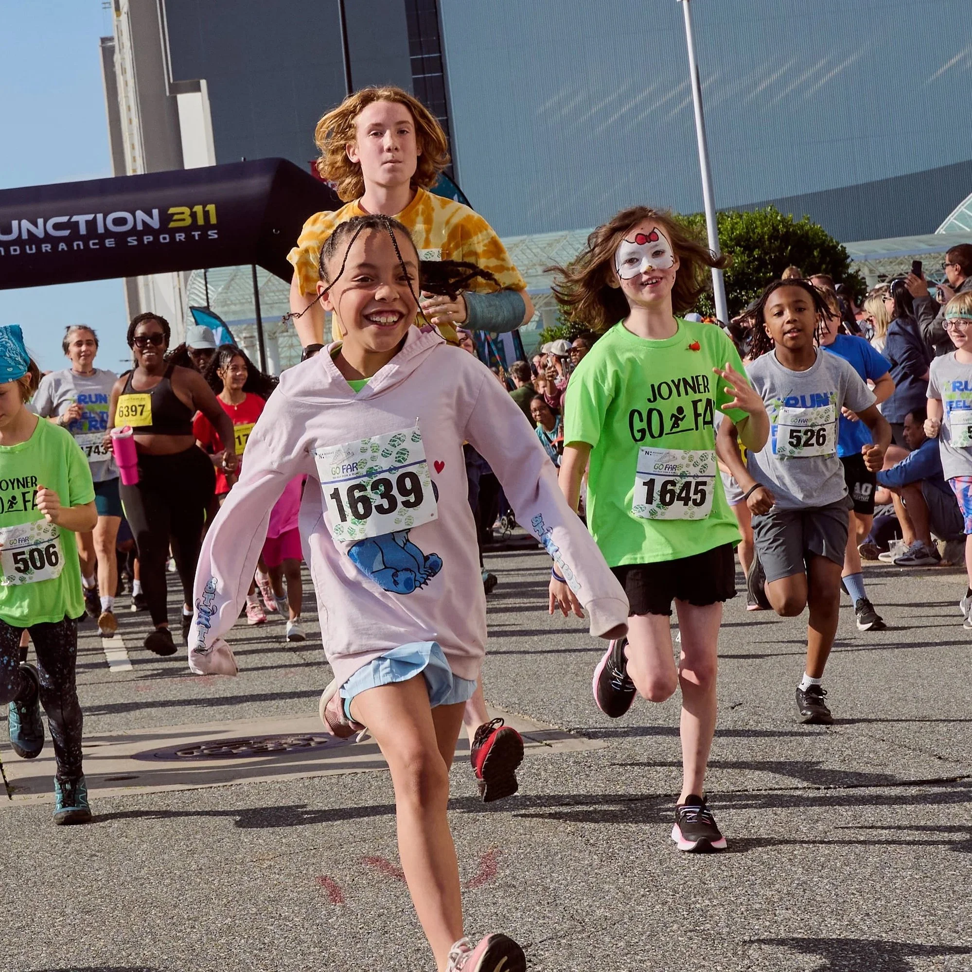 Children participating in a running event outdoors, wearing race bibs and athletic clothing, with a crowd and modern buildings in the background.
