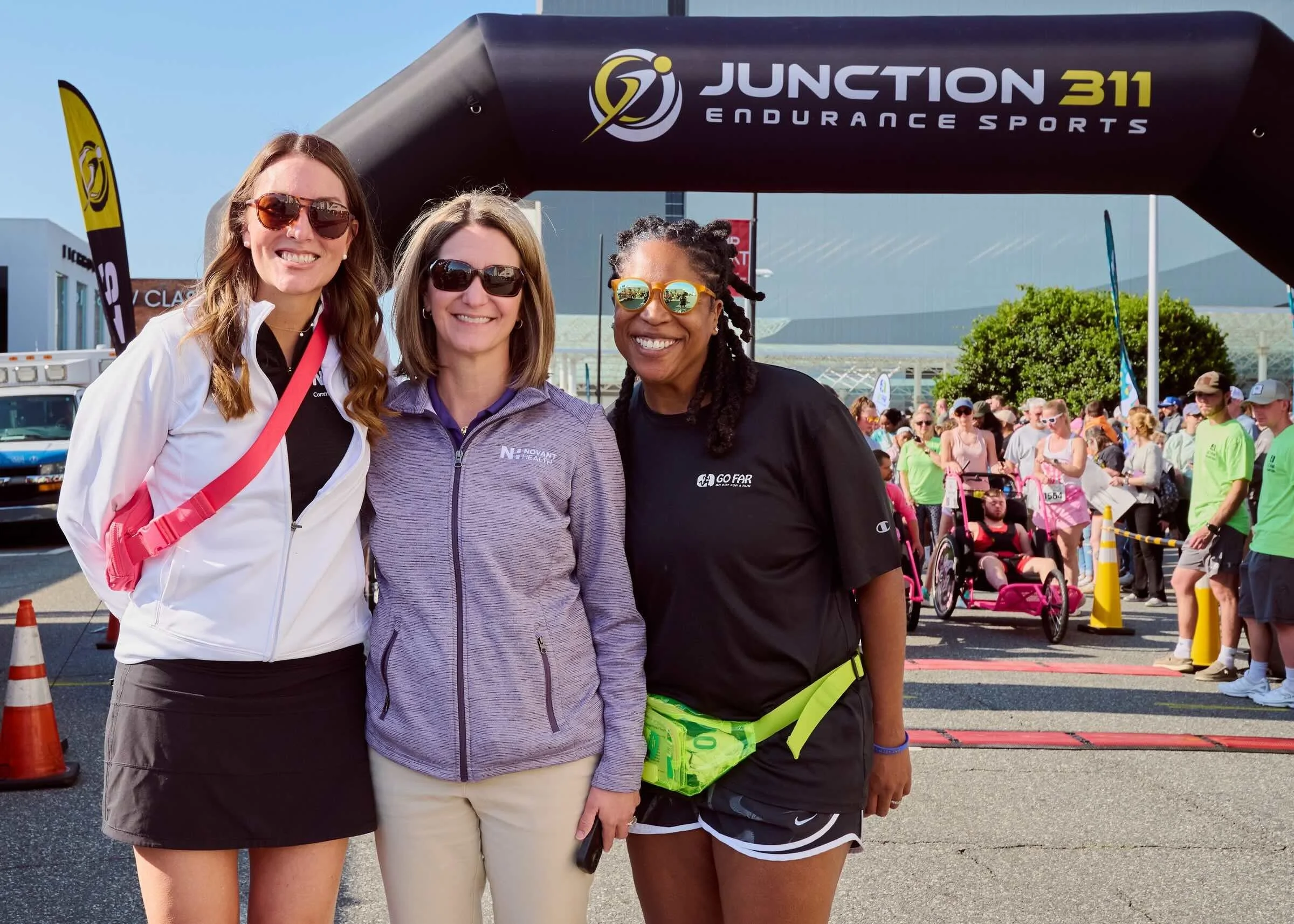 Three women standing outdoors at a running event, smiling, with a crowd, event arch, and canal in the background.