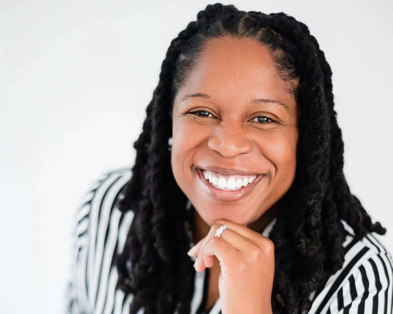 Close-up portrait of a woman smiling with her hand resting on her chin, wearing a black and white striped blazer.