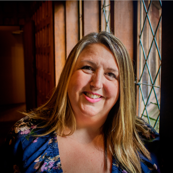 A smiling woman with blonde hair and a floral blouse sitting near a window with decorative ironwork, inside a wooden building.