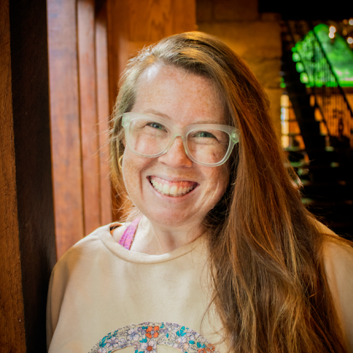 Woman with long red hair, glasses, and a beige sweatshirt smiling indoors with wooden walls and a staircase in the background.