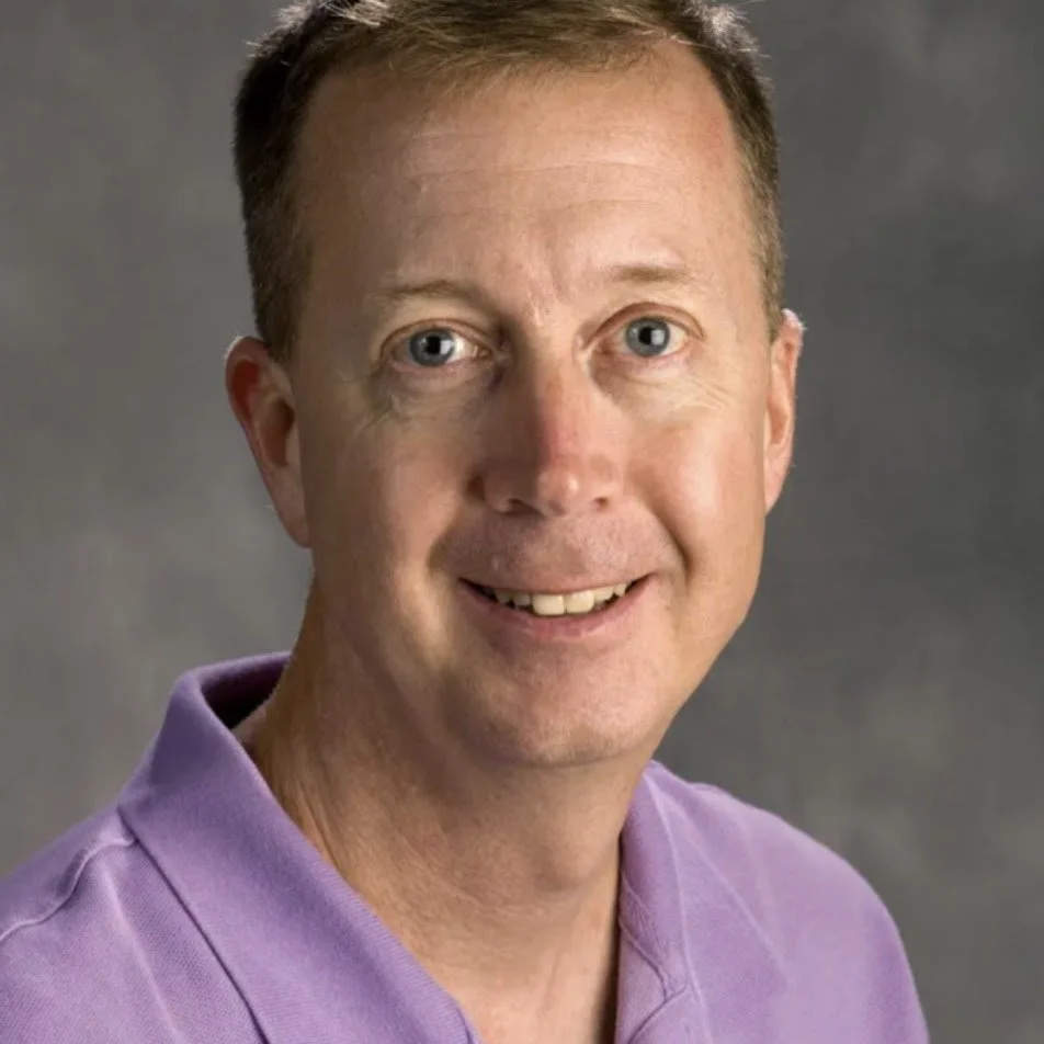 Headshot of a smiling man with short brown hair wearing a purple collared shirt against a gray background.