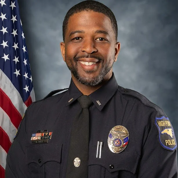 Photograph of a smiling African American police officer in uniform standing in front of an American flag with a gray background.