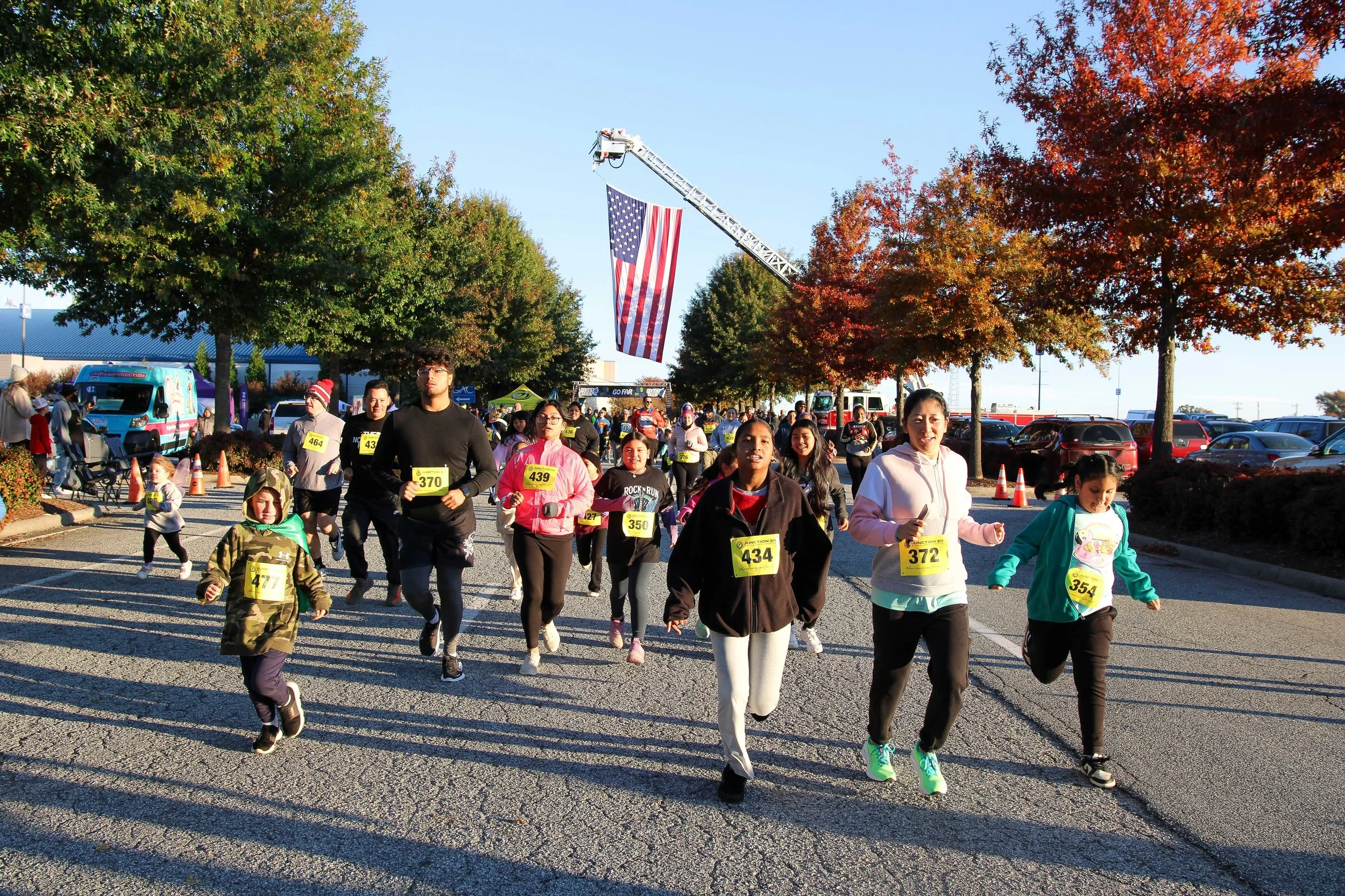 People participating in a community race or fun run outdoors on a clear day, with colorful trees, parked cars, and a large American flag hanging from a crane in the background.
