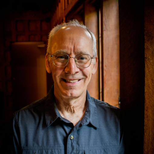 A smiling older man with glasses and gray hair, wearing a dark shirt, standing near a wooden window in a warmly lit room.
