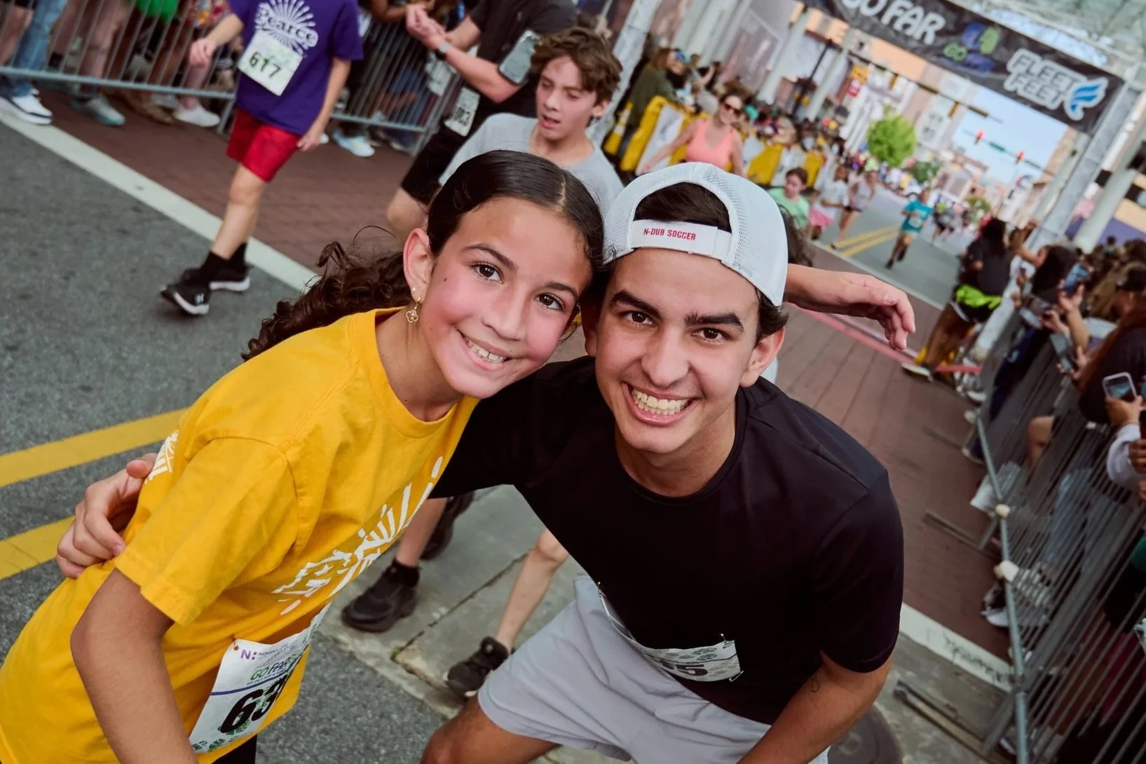 Two smiling young people, a girl with dark hair in a yellow t-shirt and a guy with a cap and black t-shirt, posing for a photo at a public event or race, with a crowd and event finish line backdrop in the background.