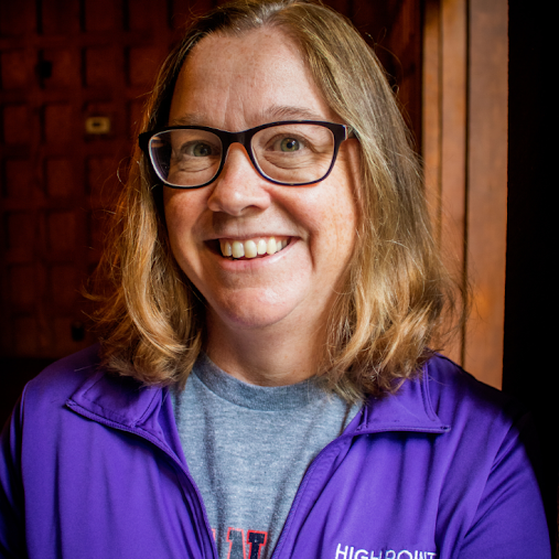 A woman with shoulder-length blonde hair and glasses smiling at the camera, wearing a purple jacket over a gray shirt, standing indoors with a wooden wall background.