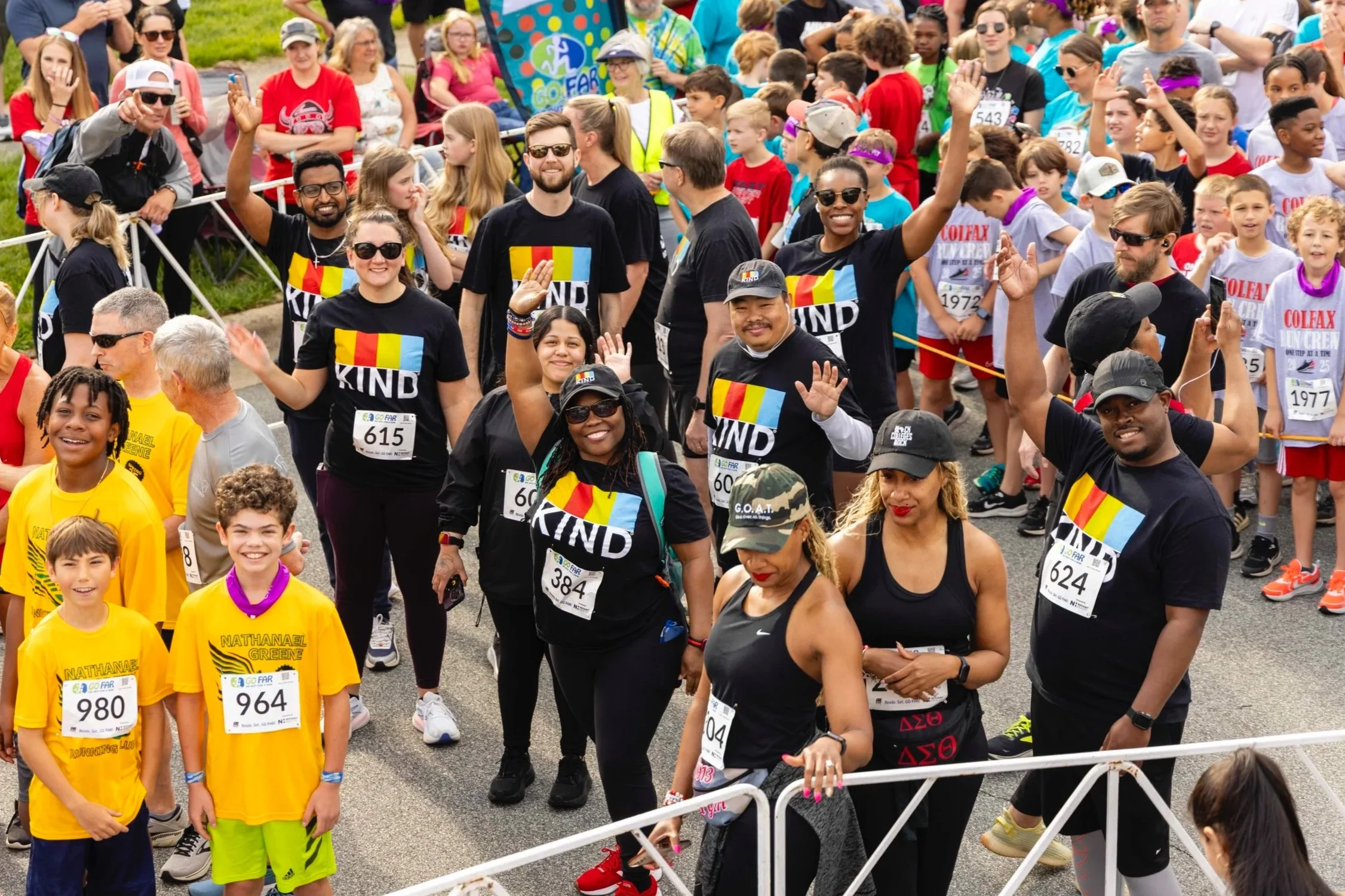 A diverse group of people participating in or spectating at a race event, with some volunteers in black shirts reading 'KIND' and military-style caps, some children and adults wearing race bibs, standing behind a white barricade on a sunny day.