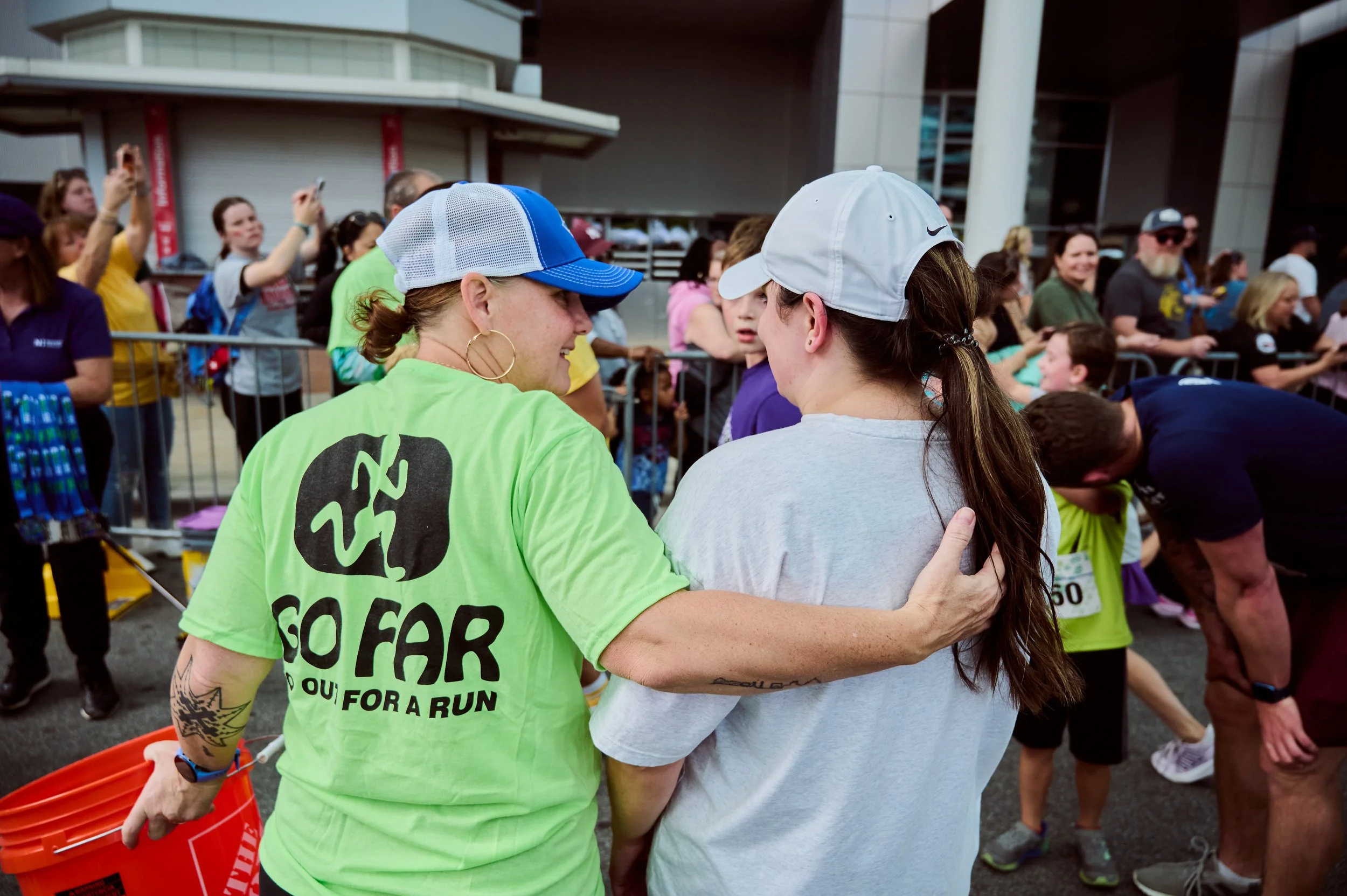Two women hugging at a race event, surrounded by other participants and spectators.