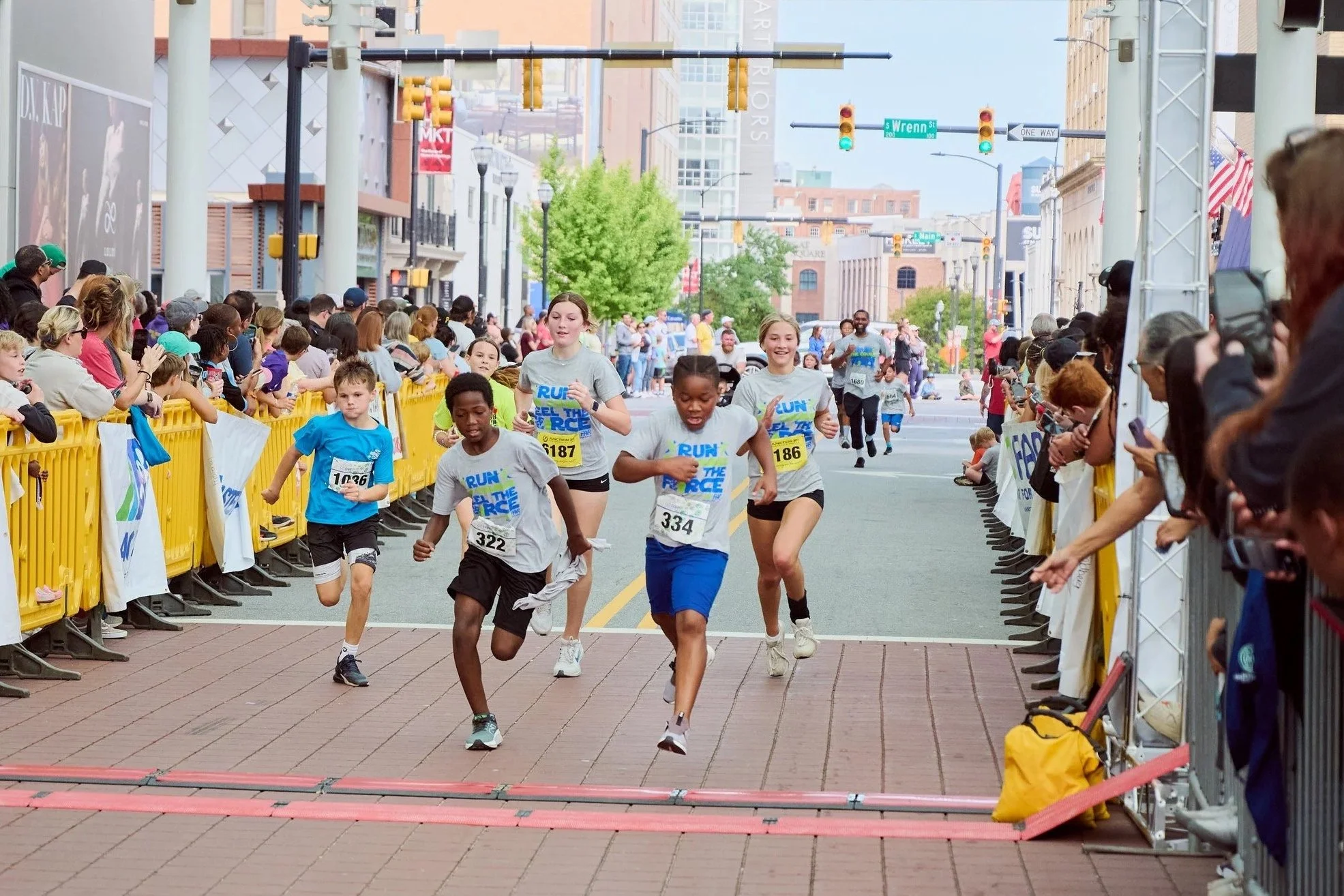 Children running in a street race with a crowd of spectators on both sides of the street, buildings in the background, yellow barriers, and traffic lights overhead.