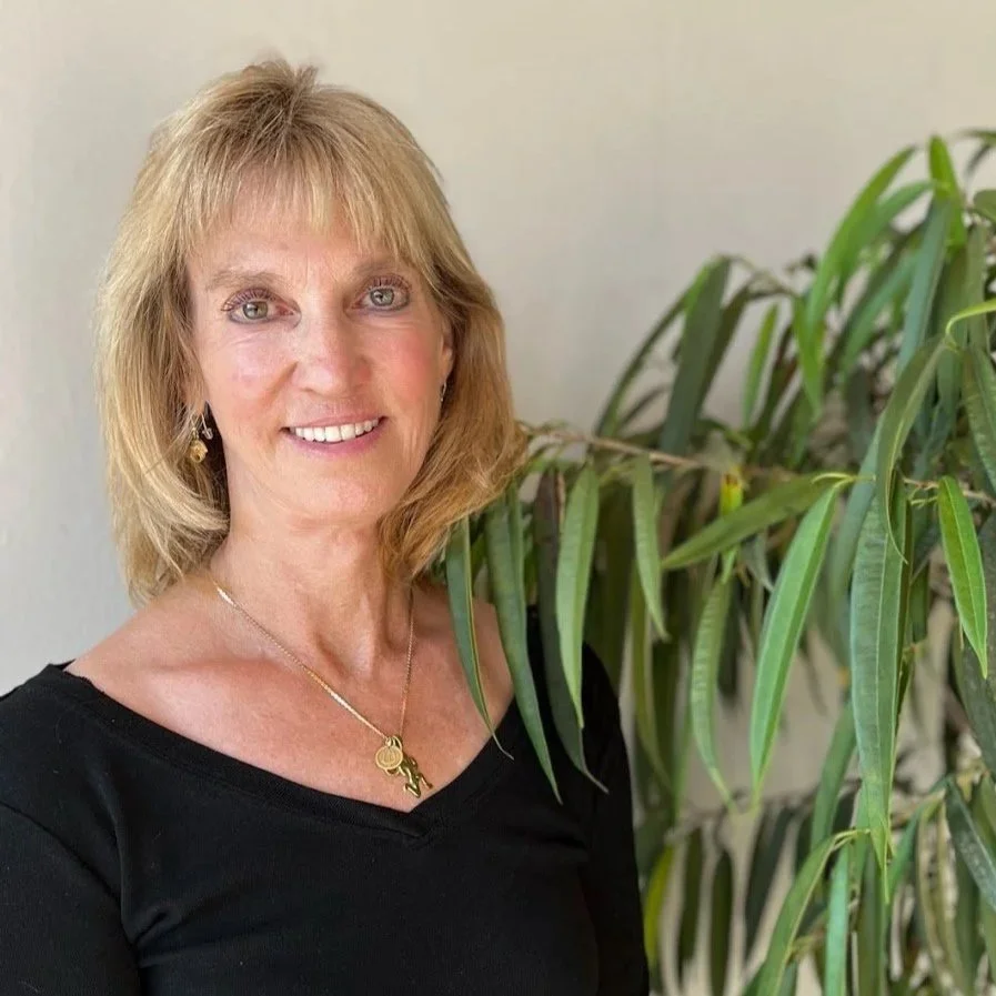 A smiling middle-aged woman with shoulder-length blonde hair, wearing a black top, gold jewelry, standing next to a large green plant with long leaves in an indoor setting.