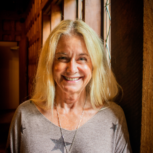 Smiling woman with shoulder-length blond hair, wearing a gray shirt with star patterns, standing indoors near a wooden wall with vertical slats, natural light from a window.