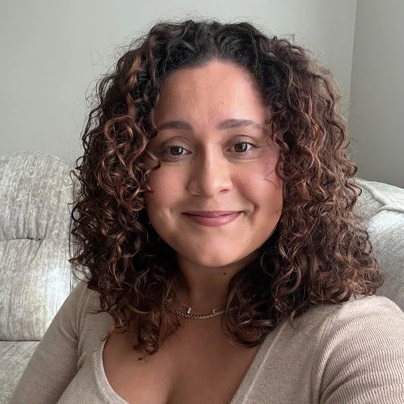A woman with curly brown hair smiling, sitting on a light-colored sofa in a room with light-colored walls.