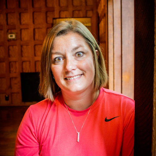 A woman with shoulder-length brown hair, wearing a pink Nike athletic shirt and a silver necklace, smiling in an indoor setting with wooden walls.