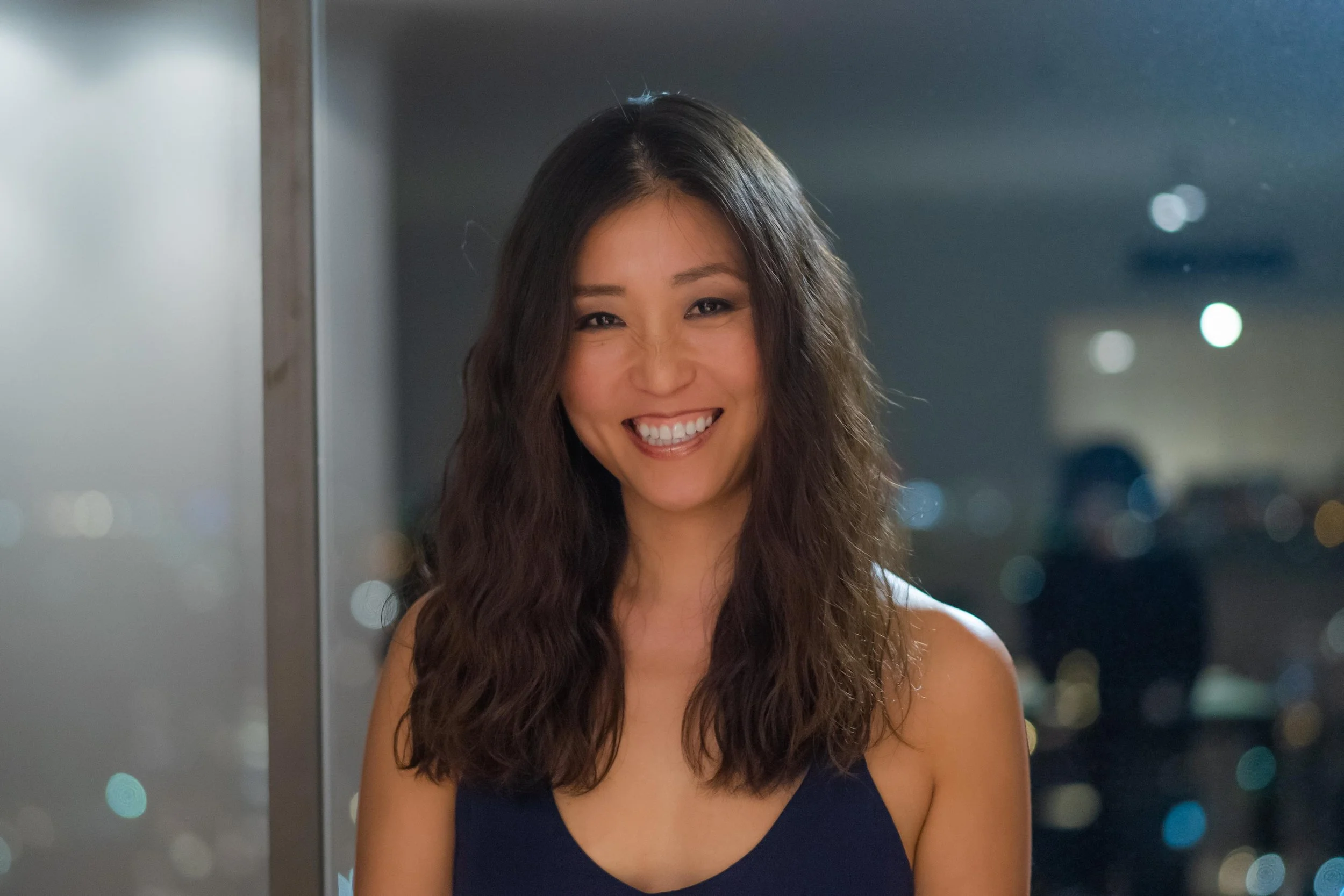 A woman with long, wavy hair smiling at the camera, wearing a dark sleeveless top, with a blurred cityscape or interior background featuring bright lights.