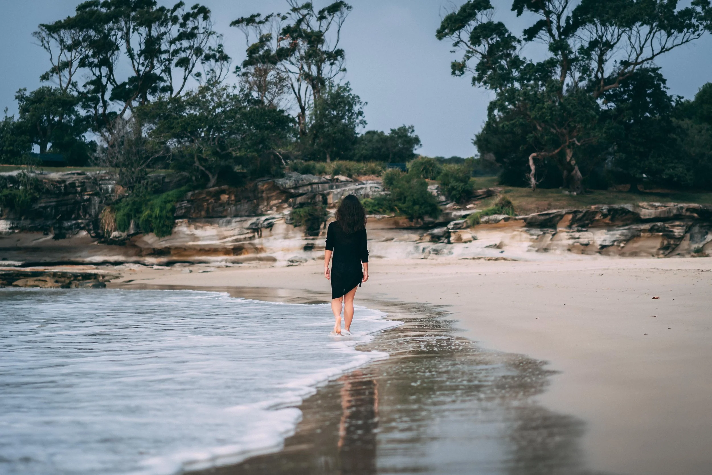 A woman in a black dress walking barefoot on a beach with her back to the camera, viewing the ocean and rocky shoreline with tall trees in the background during evening.