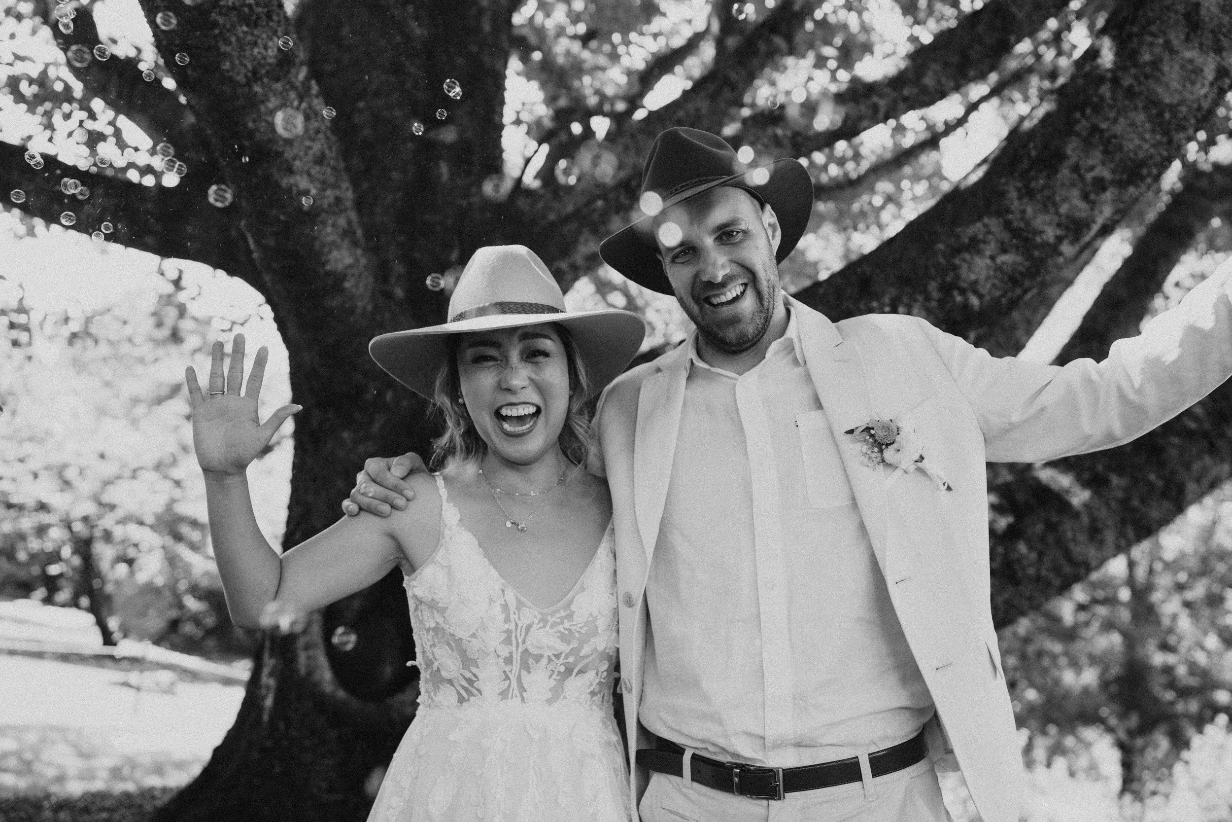Black-and-white photo of a smiling bride and groom in wedding attire standing outdoors under a large tree, waving, representing a joyful, committed, and lasting relationship.
