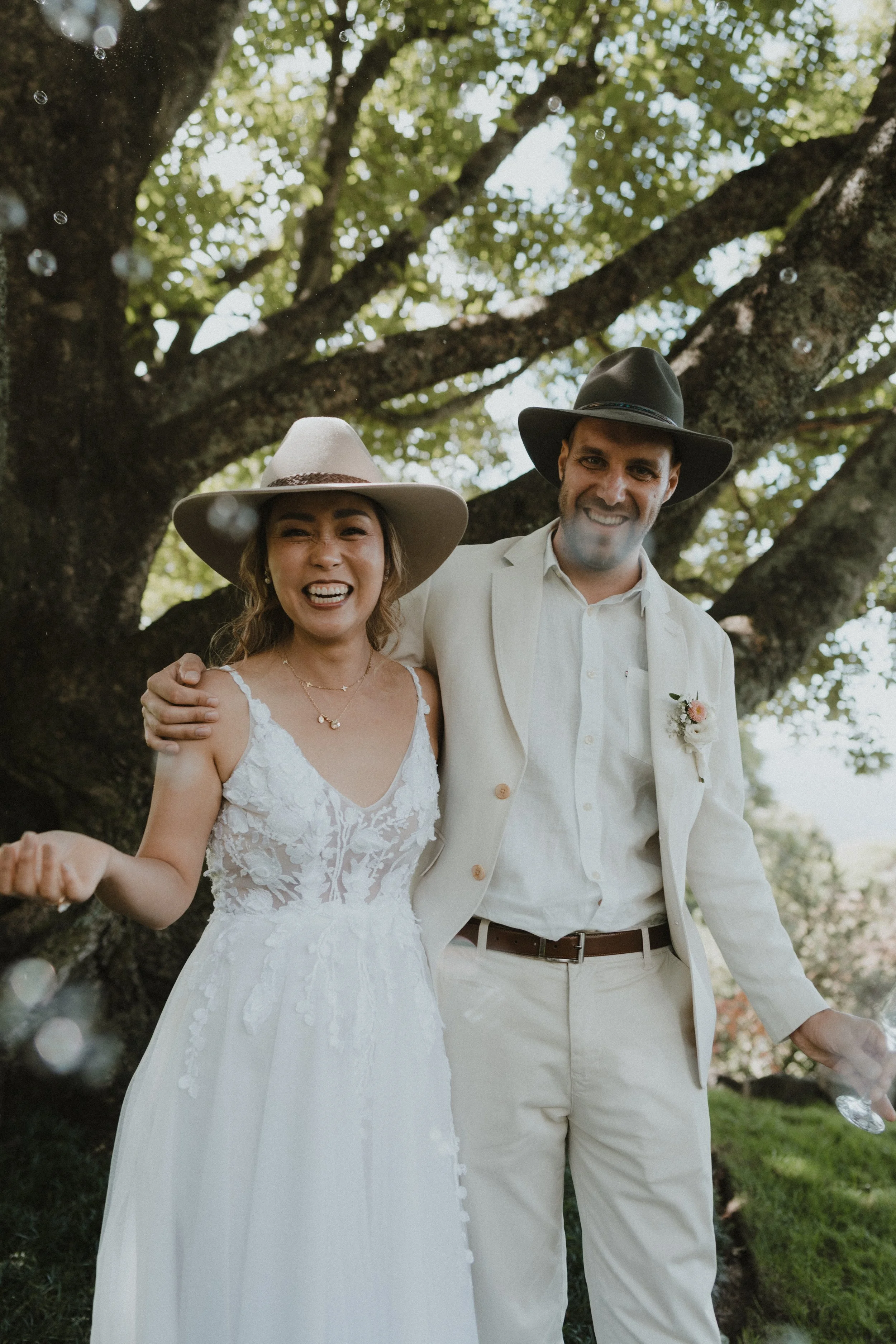 A couple dressed in wedding attire, standing outdoors under a large tree, smiling and celebrating their wedding after discovering inner clarity through personal development through inner clarity coaching.