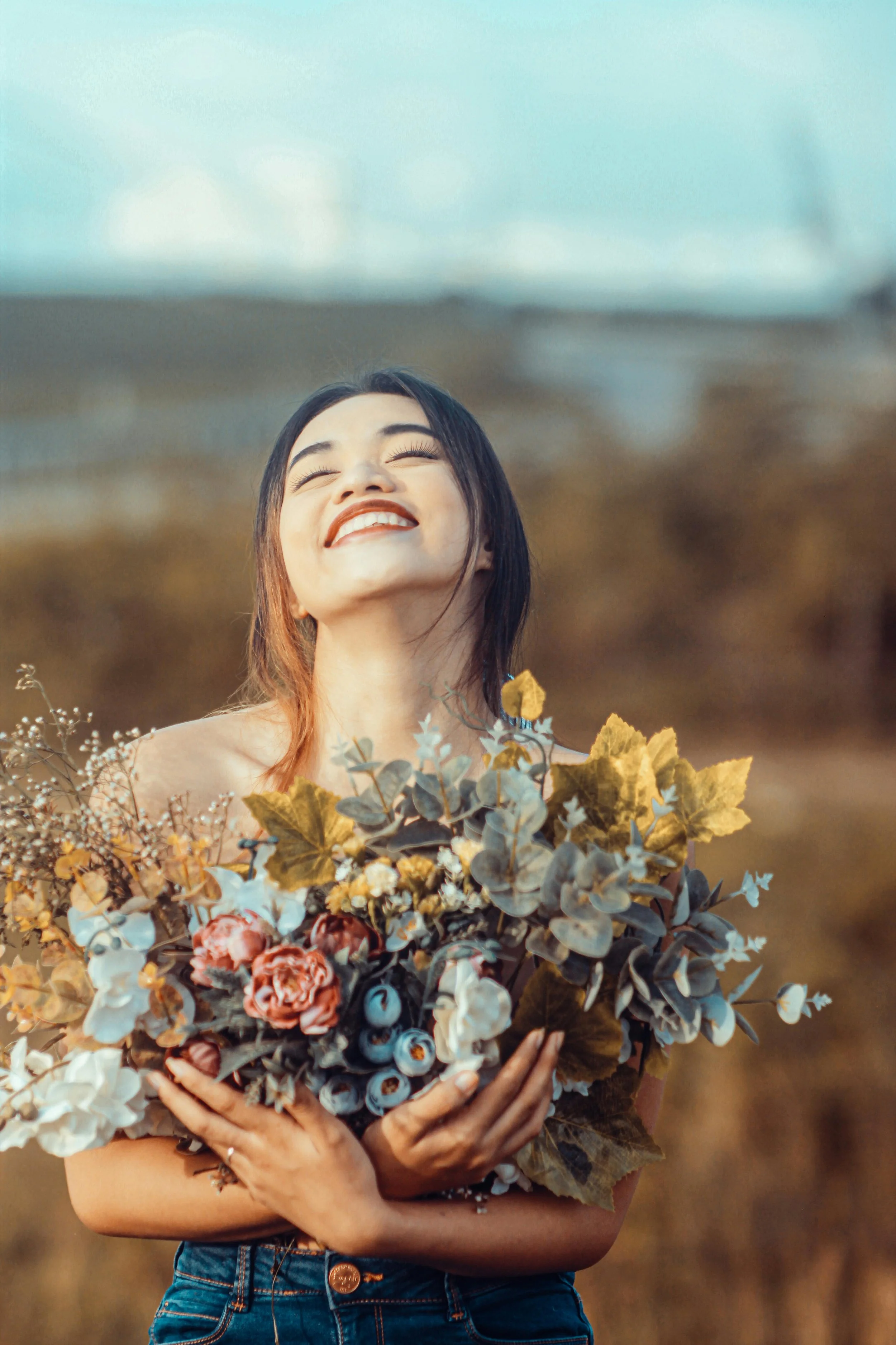A woman standing outdoors in a field, smiling with eyes closed, holding a large bouquet of flowers and foliage reflection of self love through inner clarity living