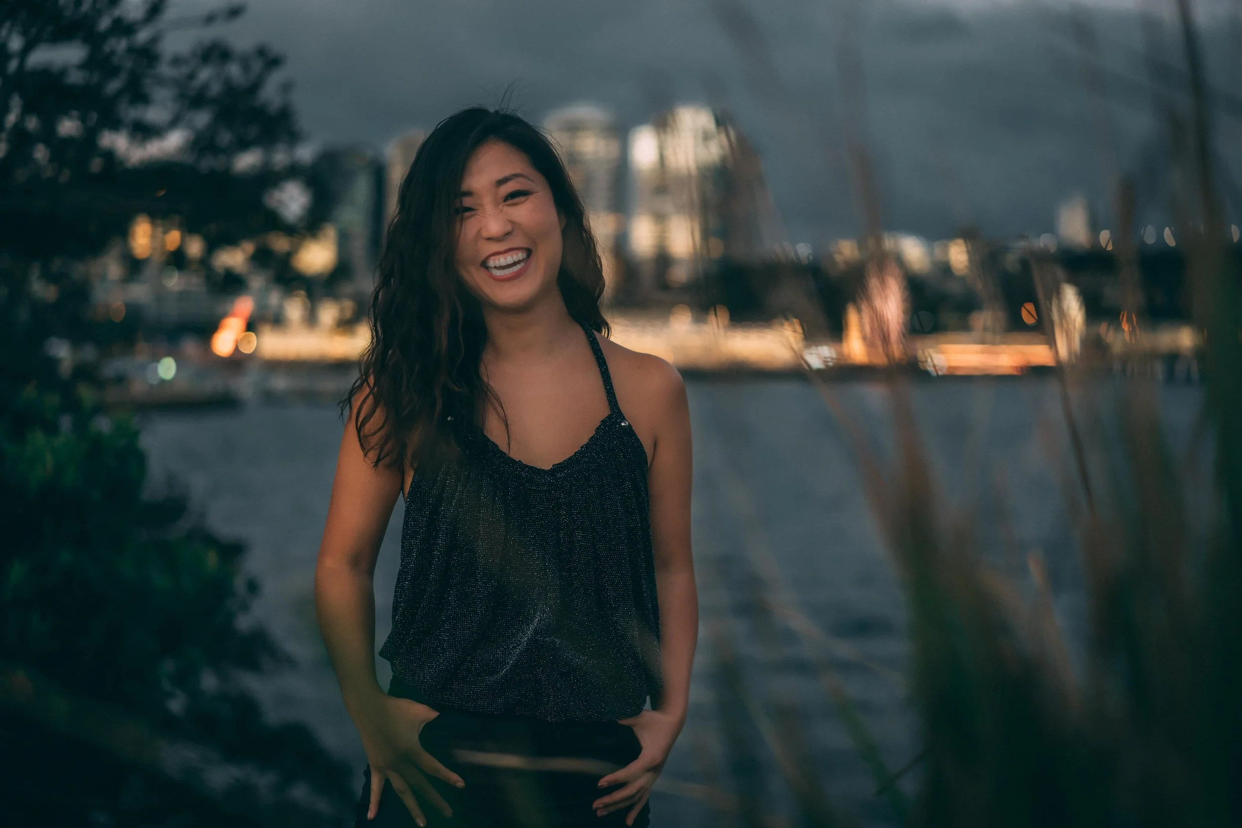 Smiling confident woman with long dark hair standing outdoors at sunset with a cityscape and trees, representing clarity, confidence, and independence for women over 40.