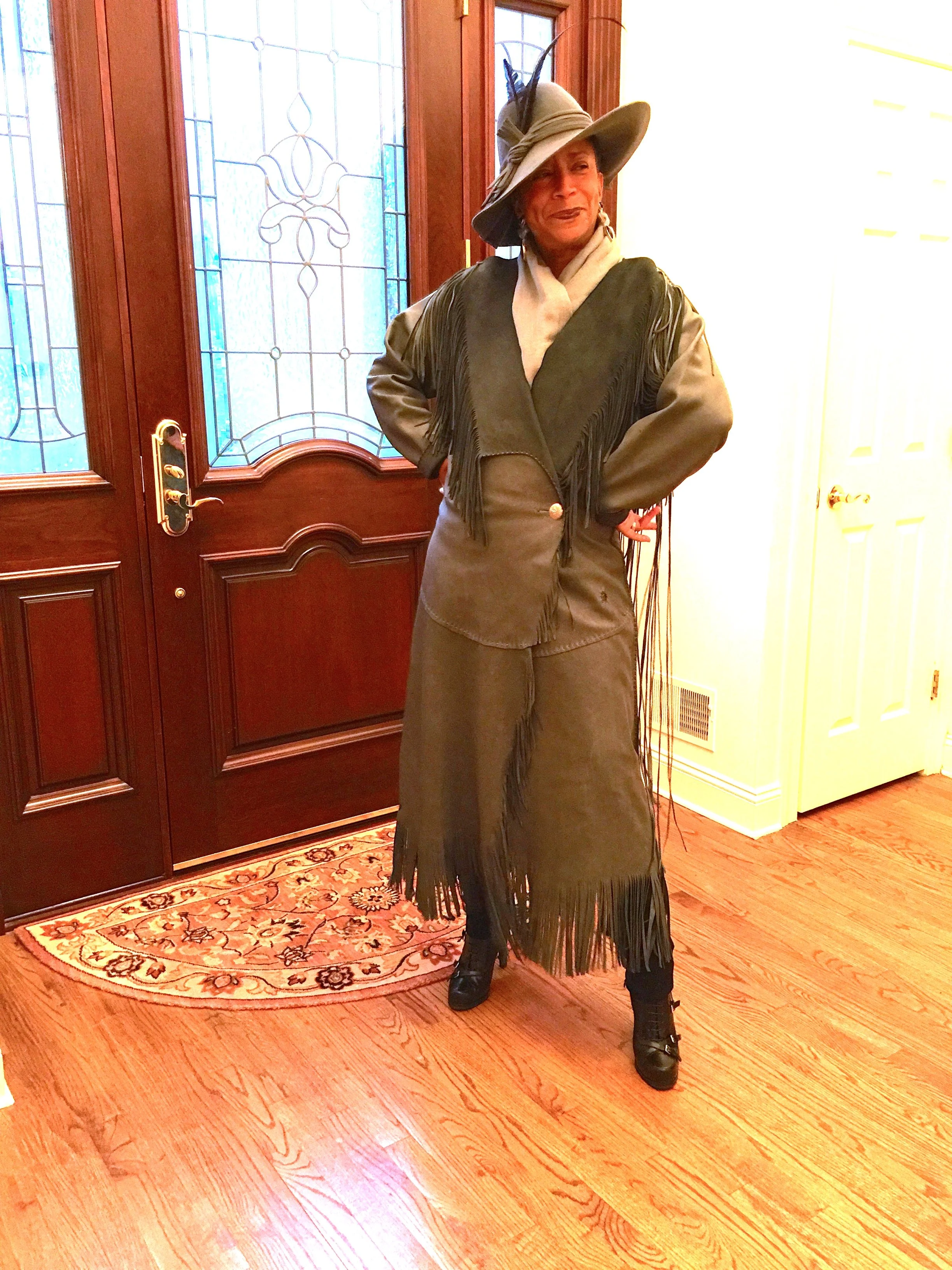 A woman standing indoors wearing a large tan hat with black feathers and a wide brim, a fringed suede jacket, a long skirt, and black heeled boots. She is standing in front of a wooden door with stained glass panels, on a hardwood floor with a patterned rug.