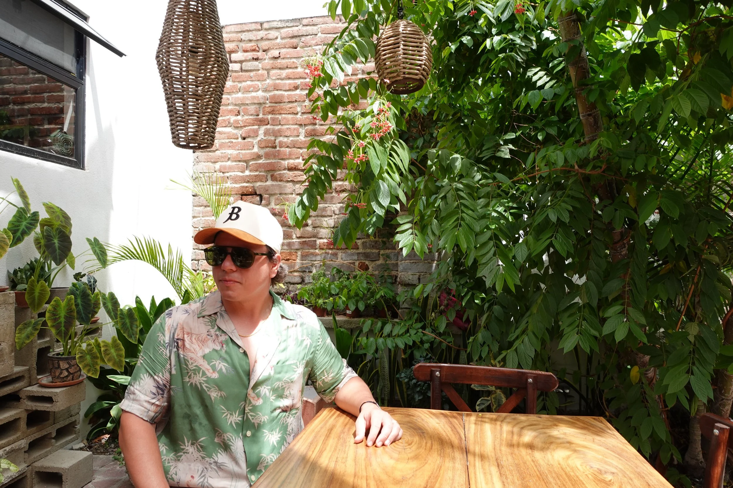 A man wearing sunglasses, a white cap with a 'B' logo, and a Hawaiian shirt, sitting at a wooden table outdoors surrounded by lush green plants and brick walls.