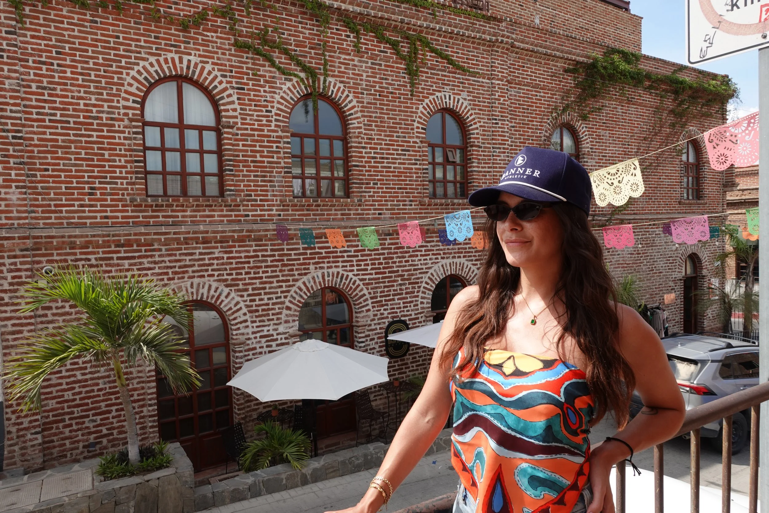 A woman wearing sunglasses, a blue cap with 'MANNER' written on it, and a colorful strapless dress with orange, blue, and yellow patterns, stands on a balcony with her hand on her hip. Behind her, there is a red brick building with arched windows, colorful papel picado banners, a white umbrella, parked cars, and some green plants.