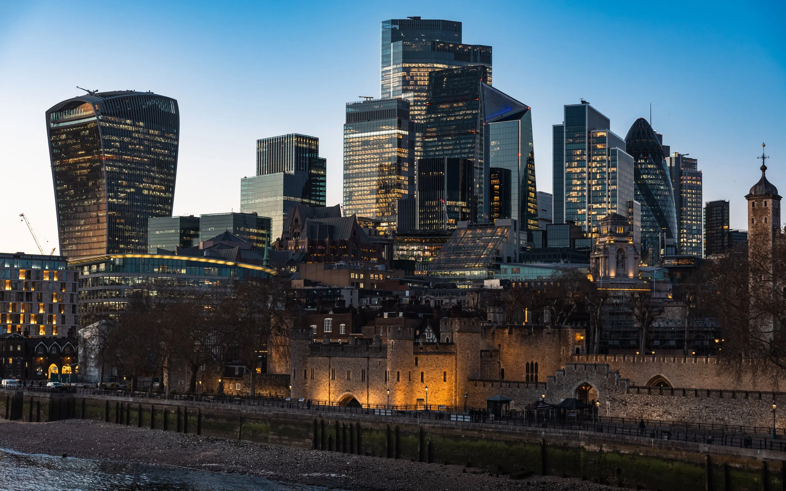 London city skyline at dusk, featuring tall modern skyscrapers and historic city walls along the river.
