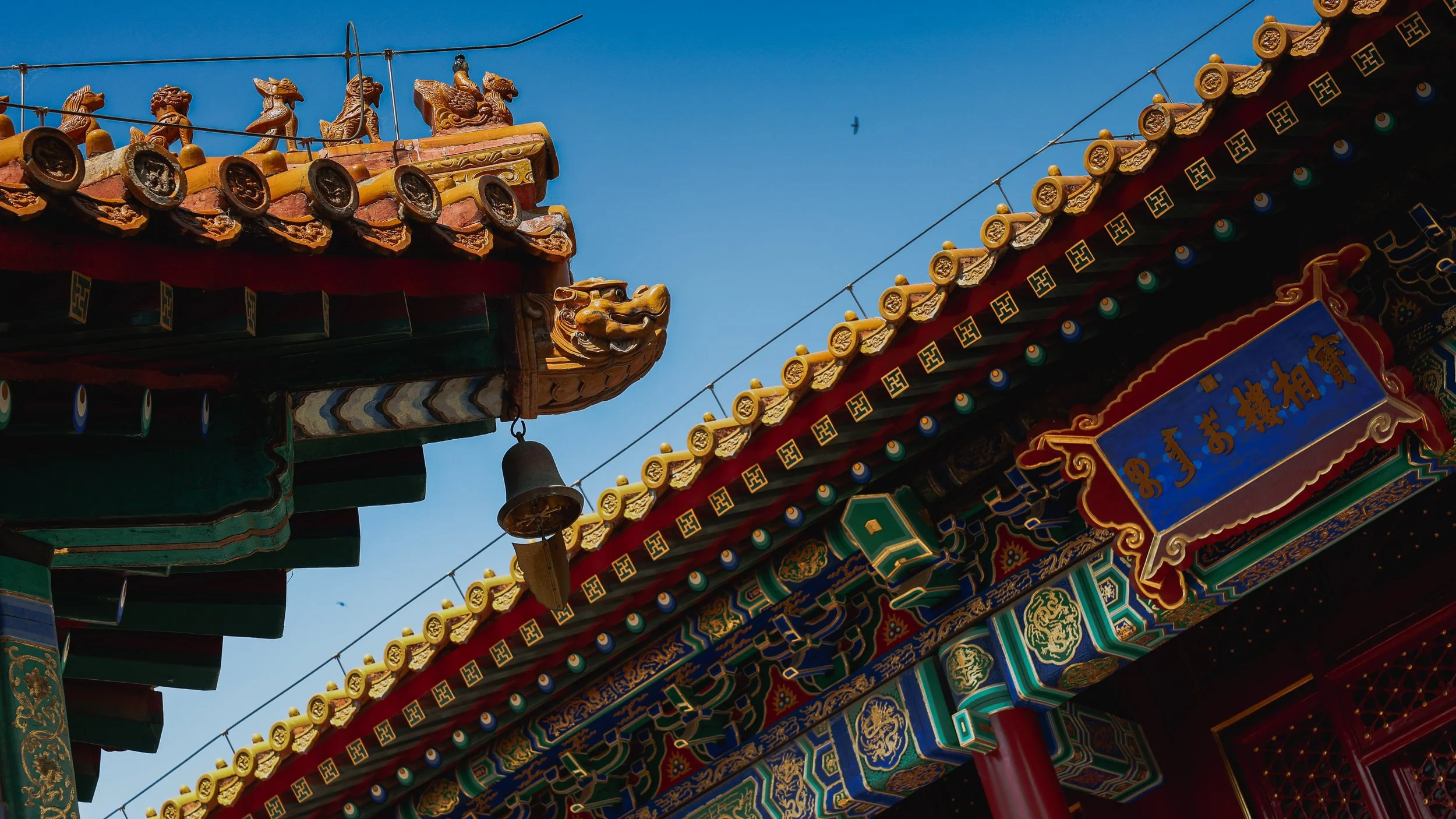 Close-up of ornate traditional Chinese temple roof with decorative figures, a bell, and a blue sky background.